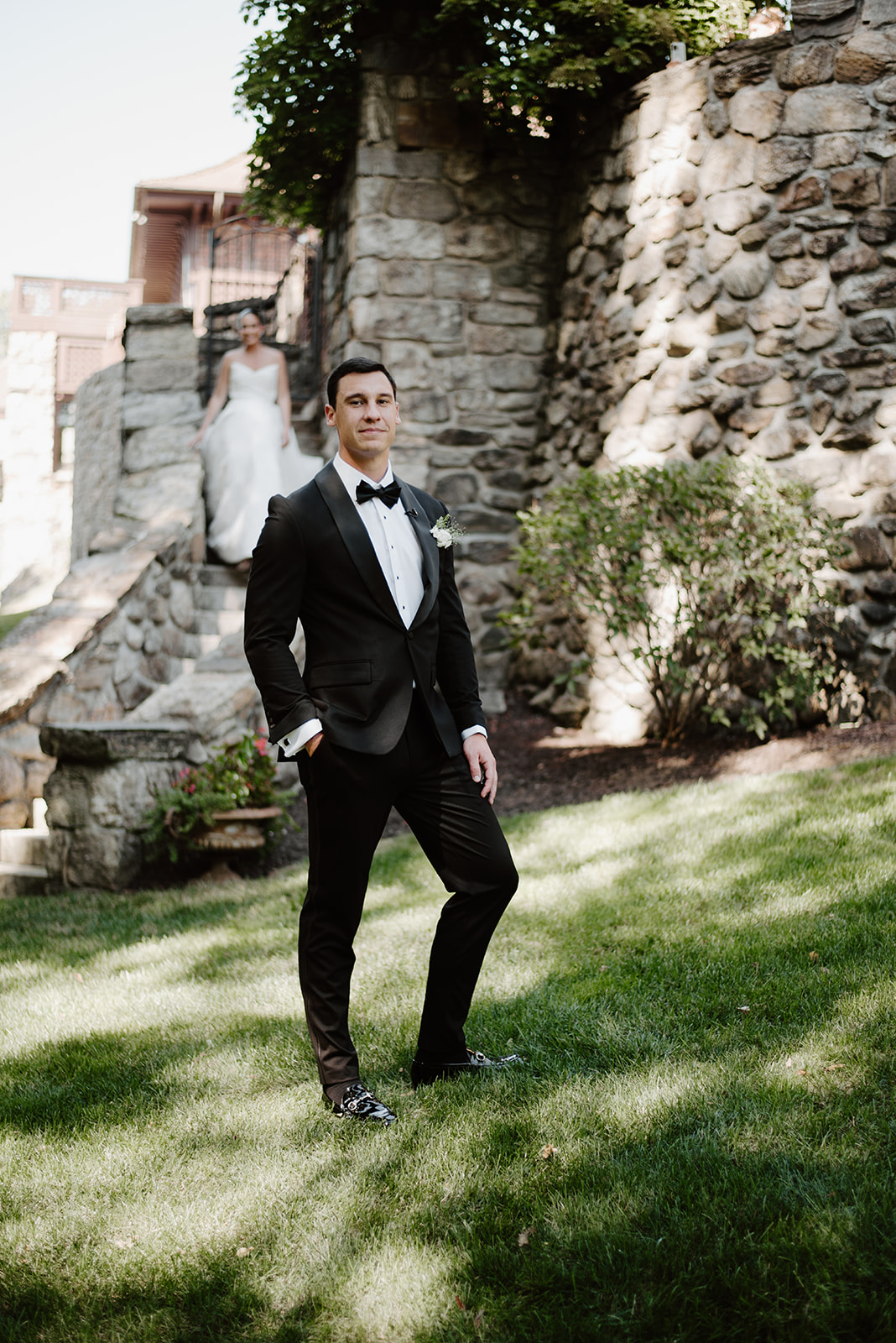Groom in a black tuxedo standing on a green lawn while the bride descends stone stairs behind him.